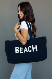 A woman with long brown hair, wearing a white t-shirt and light blue jeans, smiles while carrying the Panache Apparel Collection Beach Tote in navy blue with "BEACH" in white letters.