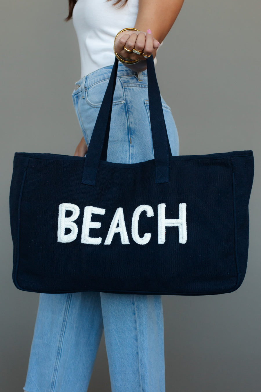 A person in a white top and blue jeans holds the Panache Apparel Collection Beach Tote, a large navy blue bag with "BEACH" printed in bold white letters, shown against a plain gray background.