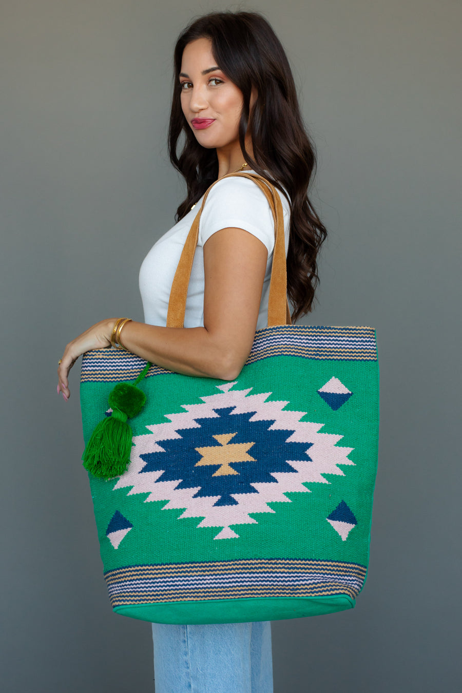 A woman with long dark hair, in a white t-shirt and light blue jeans, holds the Panache Apparel Collection Green, Light Pink & Blue Western Tote against a plain gray background.