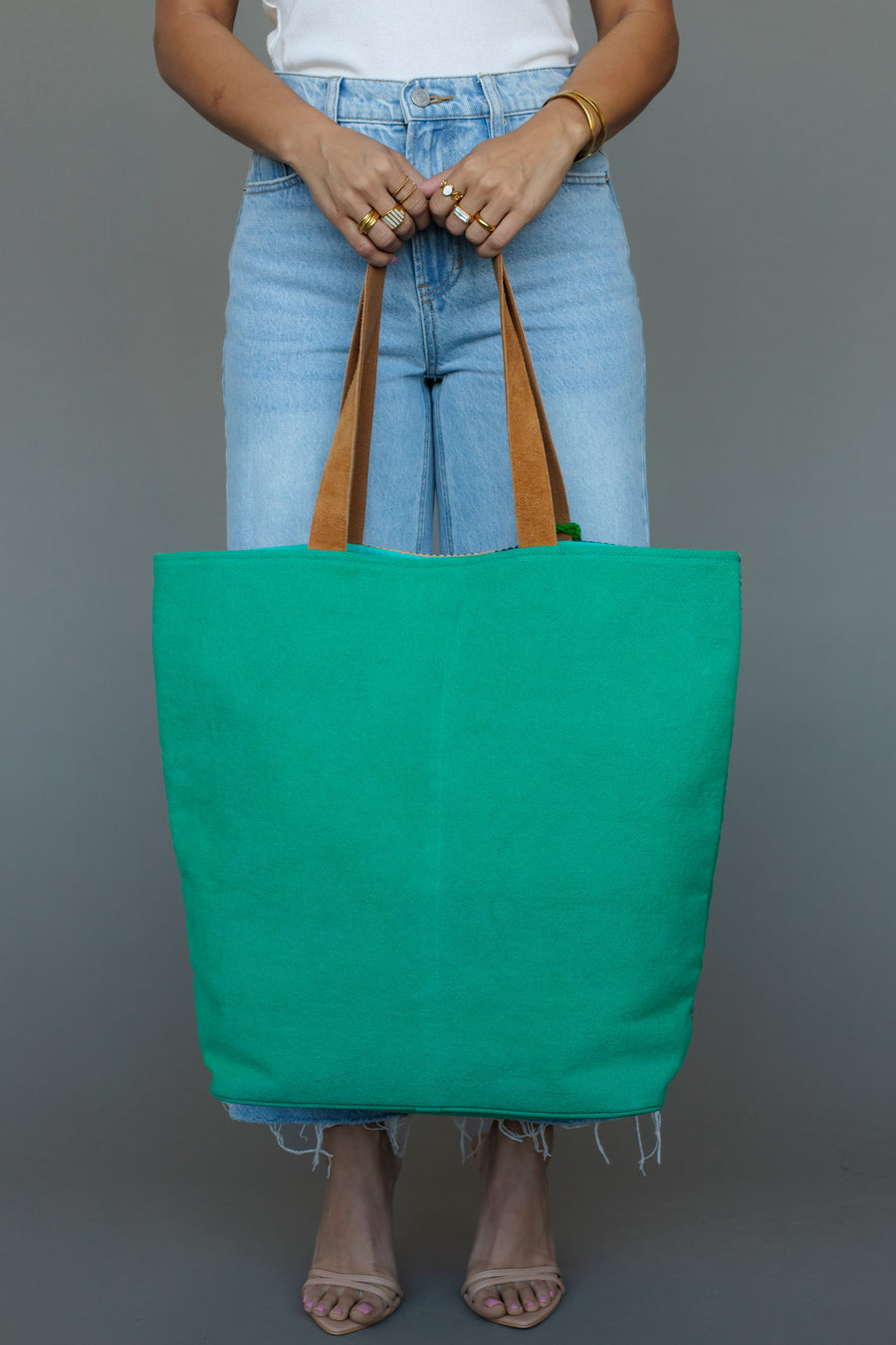 A person in light blue jeans and a white top holds the Panache Apparel Collection Green, Light Pink & Blue Western Tote, accessorized with gold rings and bracelets, standing against a plain gray background.