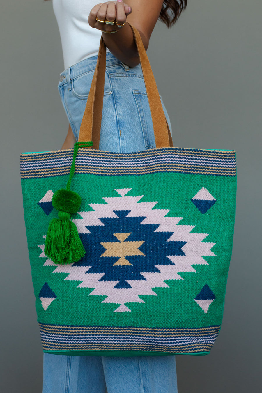 A person with long hair carries the Panache Apparel Collection Green, Light Pink & Blue Western Tote, featuring suede straps and a geometric pattern, while wearing blue jeans and a white sleeveless top.