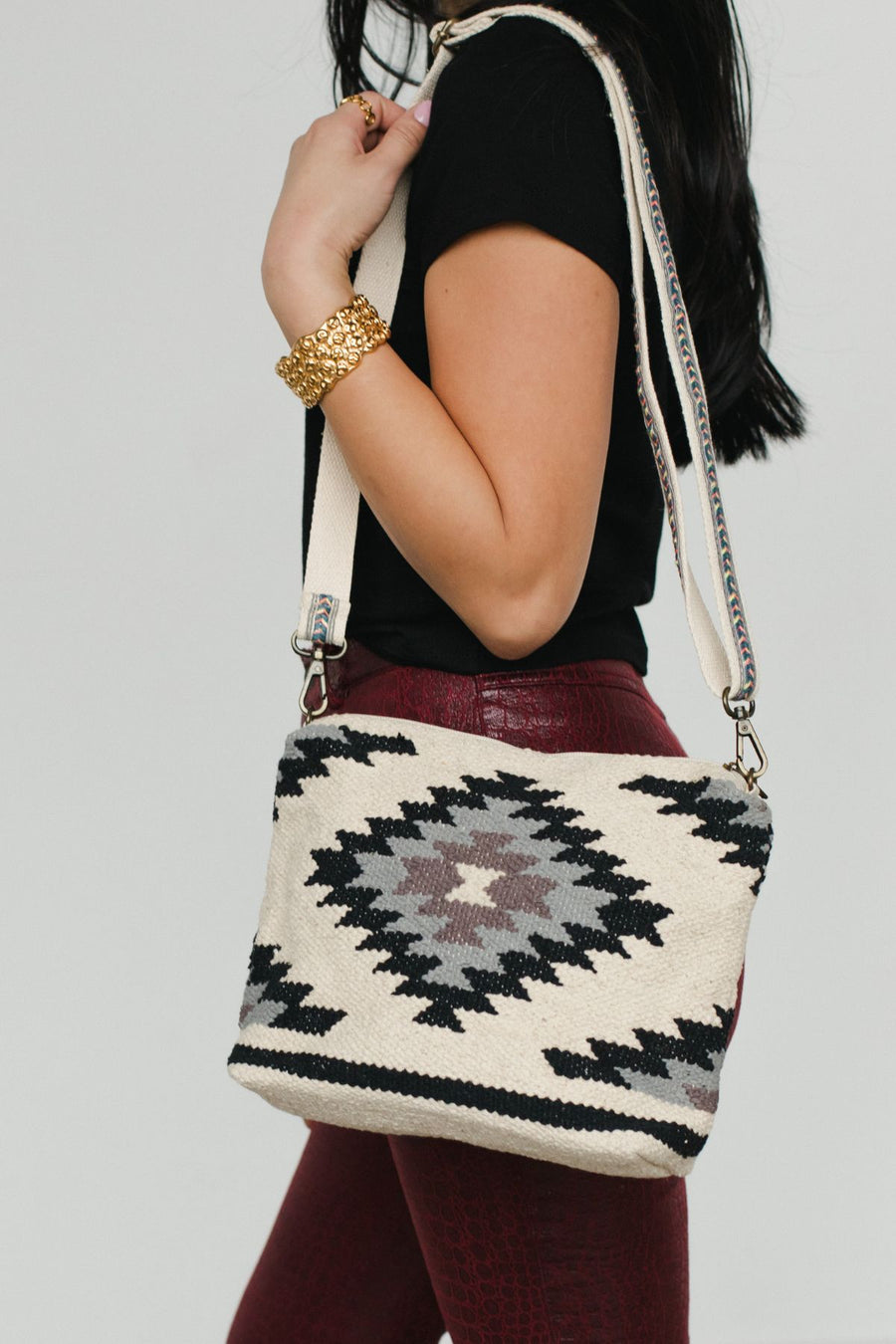 A woman with long dark hair wears a black shirt, maroon textured pants, and a gold bracelet, carrying the Panache Apparel Collection White, Black & Light Blue Western Crossbody featuring an adjustable strap and bold geometric pattern.