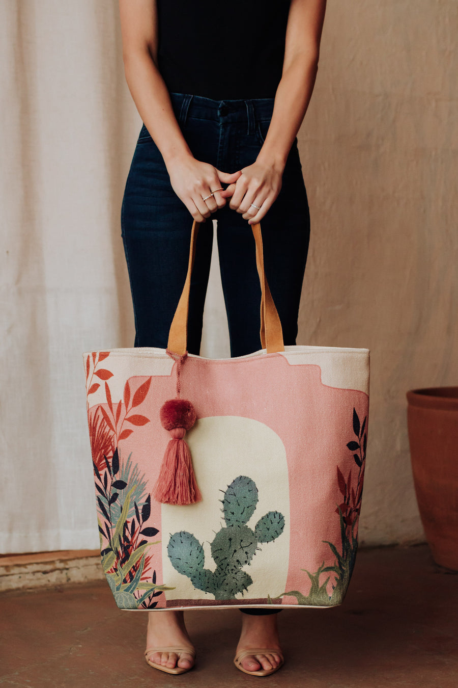 A person in a black top and blue jeans holds the Pink Cactus Landscape Tote by Panache Apparel Collection—featuring faux suede straps, colorful leaves, a cactus design, and a pink tassel. The person's face is not visible.