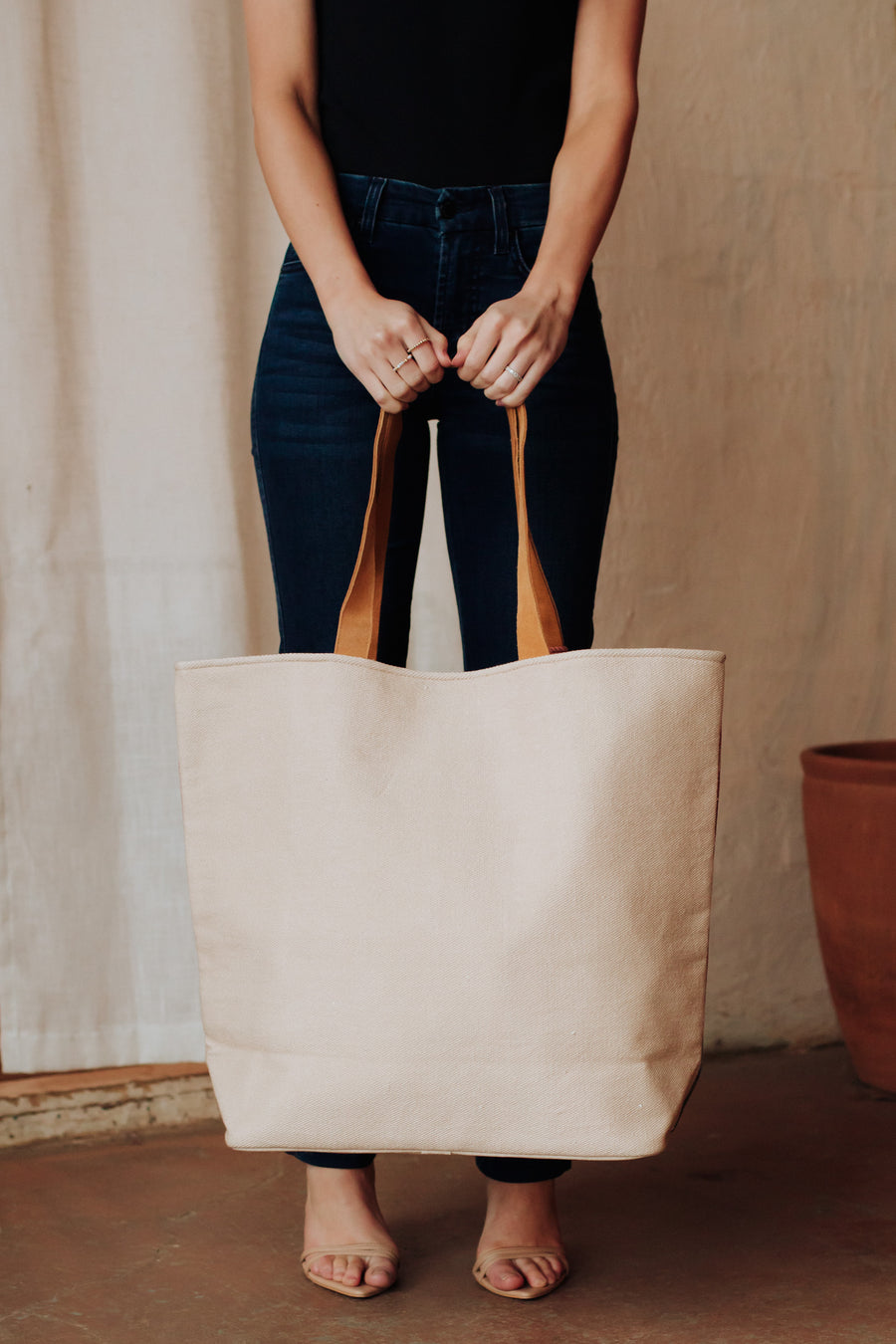 A person in dark jeans and a black top holds a large Pink Cactus Landscape Tote from Panache Apparel Collection, featuring faux suede straps, while standing indoors on a brown floor.