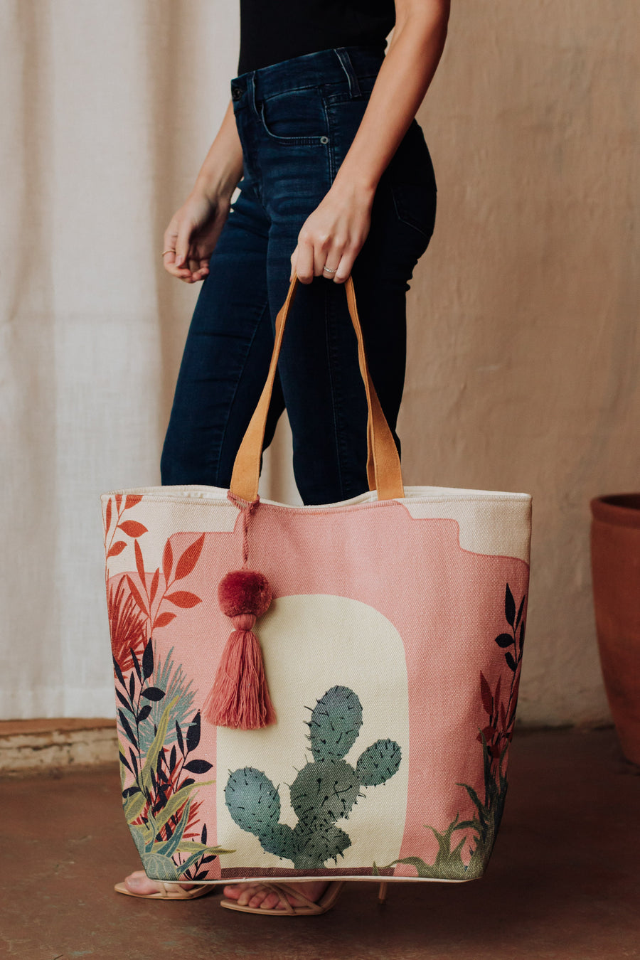 A woman in a black top and blue jeans carries the Panache Apparel Collection Pink Cactus Landscape Tote, featuring faux suede straps, a colorful plant design, and a red tassel accent for a standout accessory.