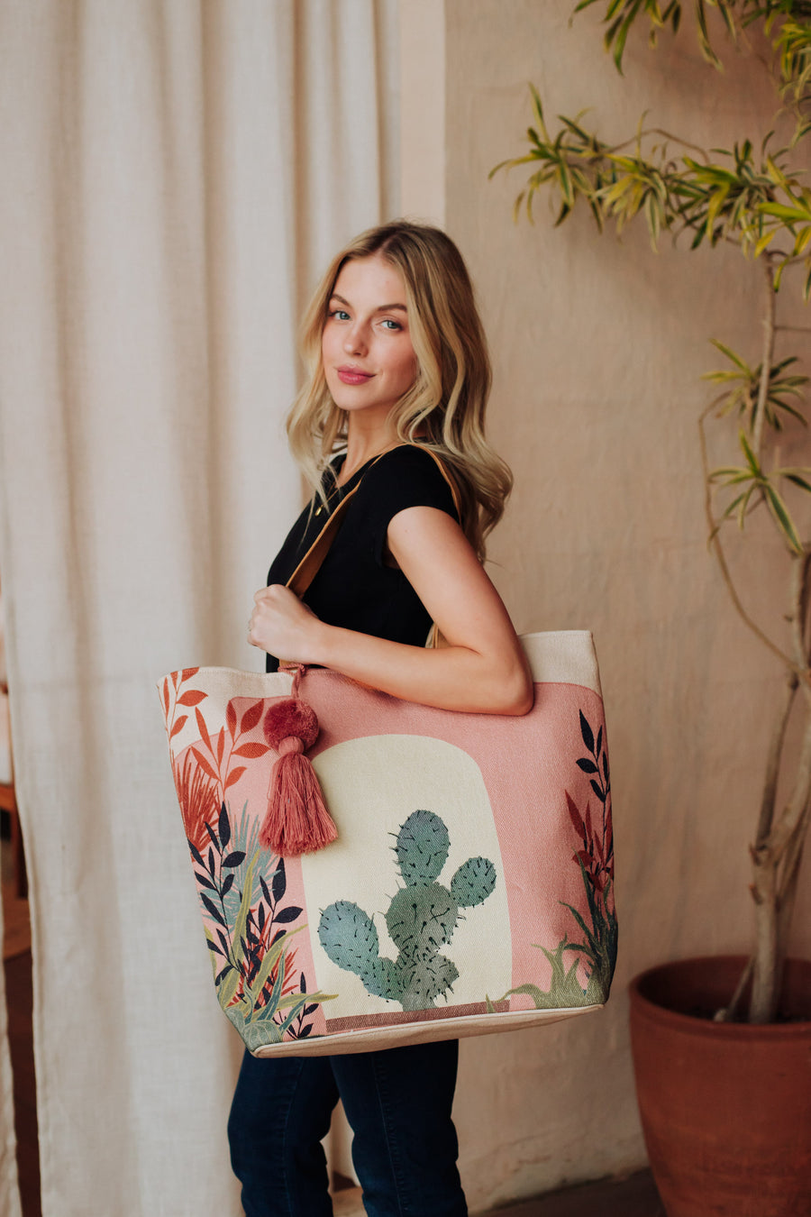 A woman with long blonde hair, in a black top and jeans, stands indoors holding the Panache Apparel Collection Pink Cactus Landscape Tote featuring faux suede straps, pink tassels, and colorful leaf designs; a potted plant sits behind her.