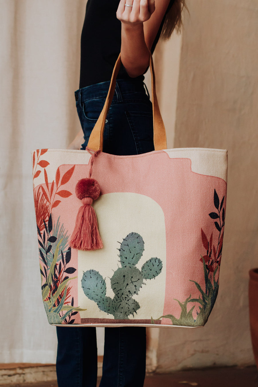 A person in a black top and jeans carries the Pink Cactus Landscape Tote by Panache Apparel Collection, featuring faux suede straps, a colorful cactus with red and green leaves, and a pink tassel decoration.