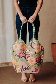 A person in a black top and jeans holds the Panache Apparel Collection Green & Multicolored Floral Tote, featuring pink, orange, and purple floral embroidery with green handles. They stand barefoot on a brown floor.