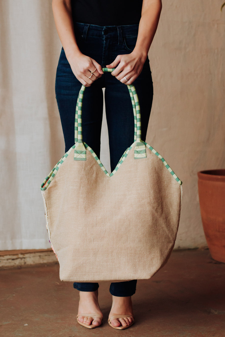 A woman in dark jeans and a black top holds the Panache Apparel Collection Green & Multicolored Floral Tote, featuring green and white checkered handles, while standing indoors on a brown floor beside a terracotta pot.