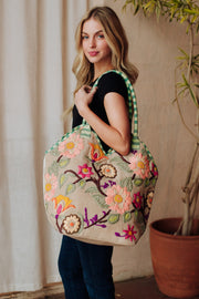 A woman with long blonde hair, in a black top and blue jeans, carries the Panache Apparel Collection’s Green & Multicolored Floral Tote, featuring green checked straps and colorful embroidery, standing indoors by a potted plant.