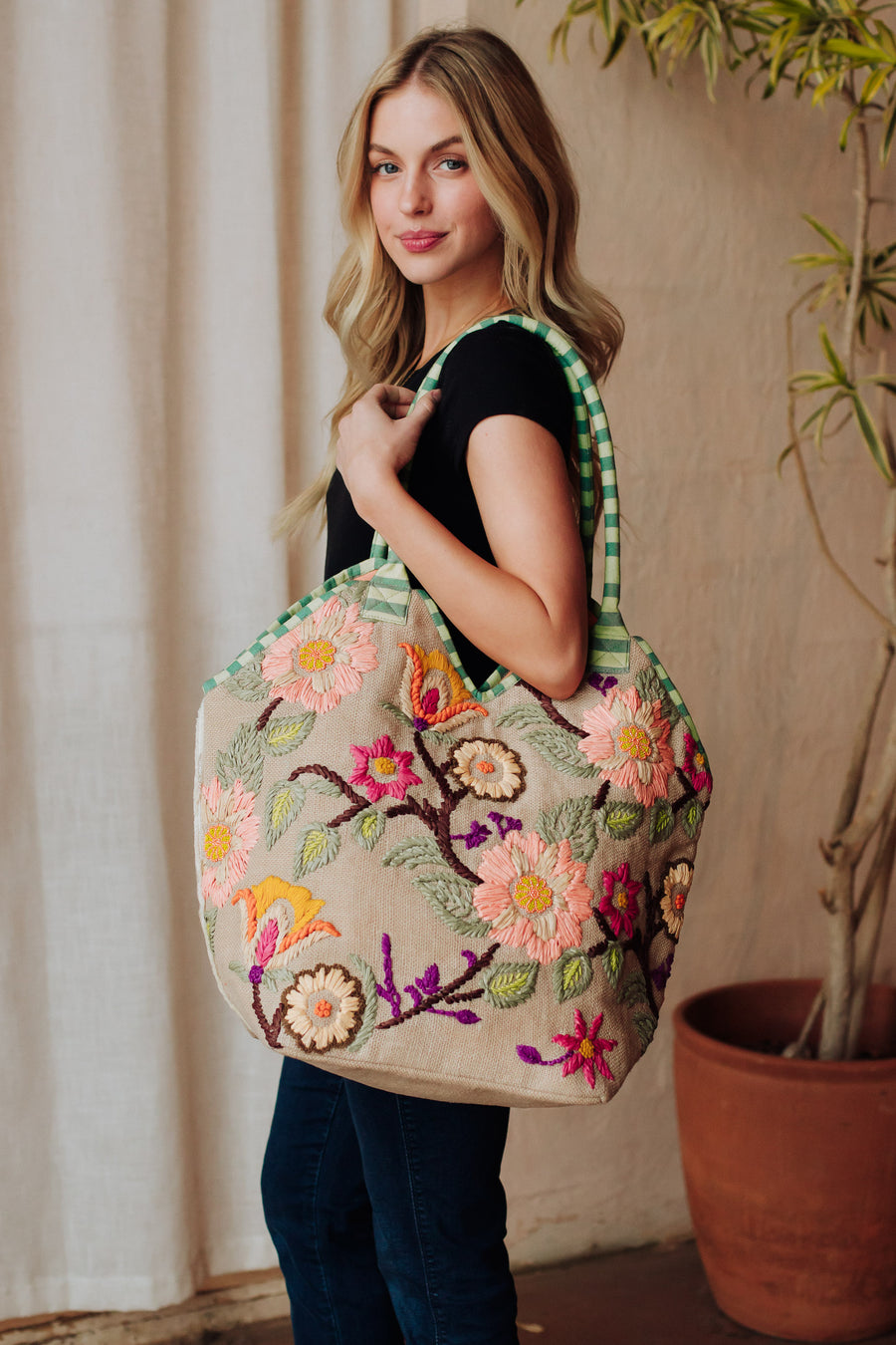 A woman with long blonde hair, in a black top and blue jeans, carries the Panache Apparel Collection’s Green & Multicolored Floral Tote, featuring green checked straps and colorful embroidery, standing indoors by a potted plant.