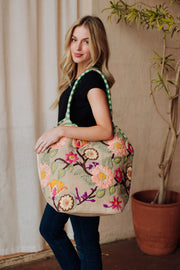 A woman with long blonde hair in a black top and jeans poses indoors, smiling with the Panache Apparel Collection Green & Multicolored Floral Tote over her shoulder. The floral-embroidered bag stands out against a backdrop featuring a potted plant.