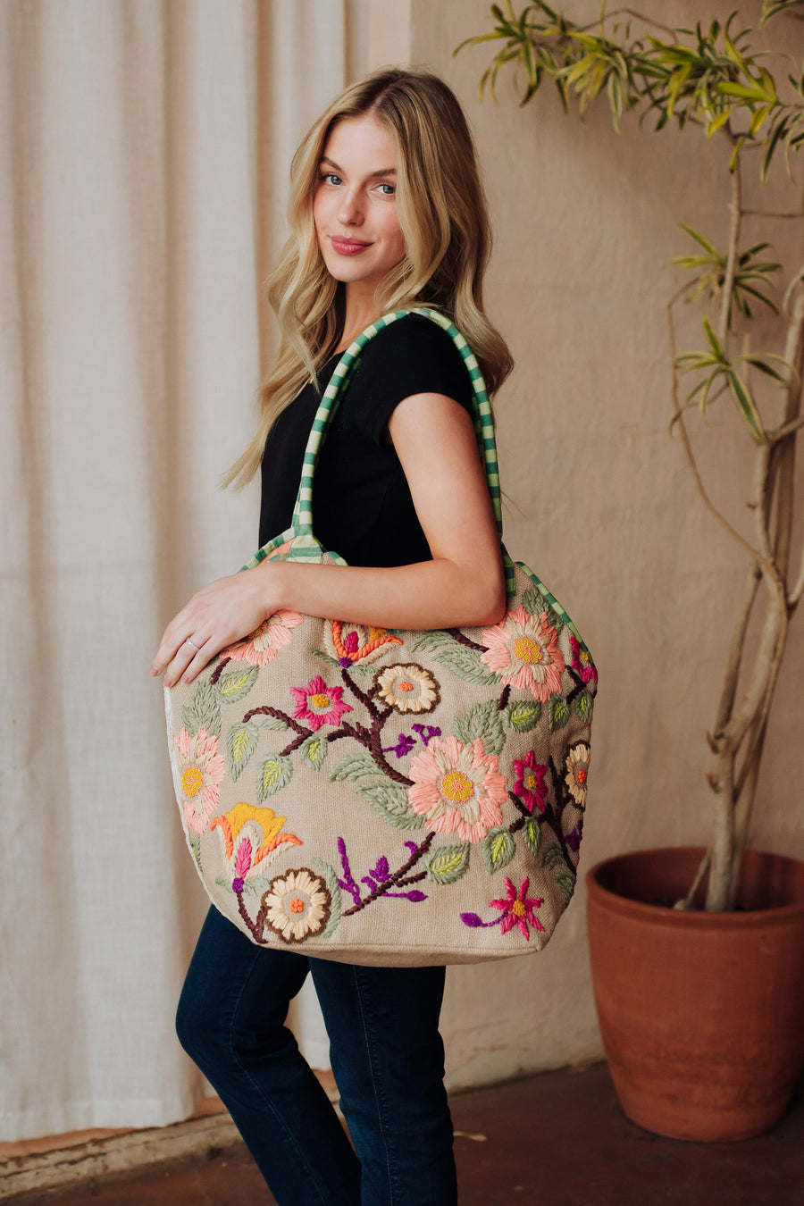 A woman with long blonde hair in a black top and jeans poses indoors, smiling with the Panache Apparel Collection Green & Multicolored Floral Tote over her shoulder. The floral-embroidered bag stands out against a backdrop featuring a potted plant.
