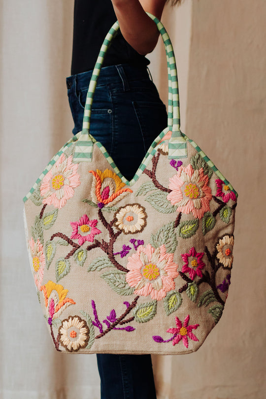 A person holds the Panache Apparel Collection Green & Multicolored Floral Tote, featuring green and white striped handles and a vibrant floral design on a beige background.