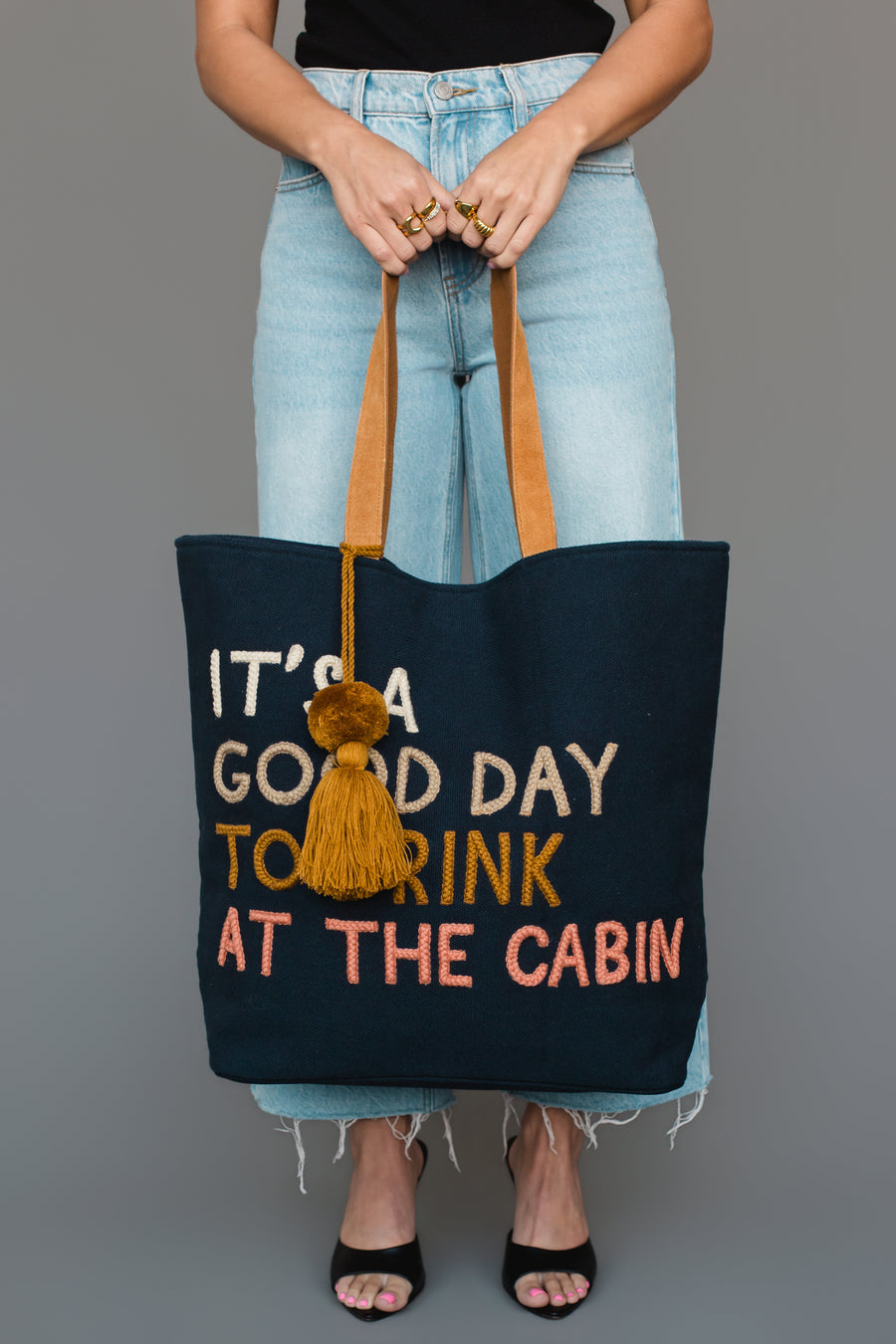 Wearing light blue jeans and black heels, a person carries the Mustard & Pink Drink At The Cabin Tote by Panache Apparel Collection, a navy bag with tan handles, a yellow tassel, and colorful lettering that reads “ITS A GOOD DAY TO DRINK AT THE CABIN.”.