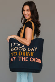 A woman with long dark hair, dressed in a black shirt and light jeans, stands against a plain gray background while smiling and holding the Mustard & Pink Drink At The Cabin Tote from Panache Apparel Collection, a large tote bag featuring the phrase “It’s a good day to drink at the cabin” in colorful letters.