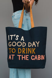 A person holds the Mustard & Pink Drink At The Cabin Tote by Panache Apparel Collection, featuring tan handles and text that reads, “ITS A GOOD DAY TO DRINK AT THE CABIN.” They are wearing light blue jeans and a black top.