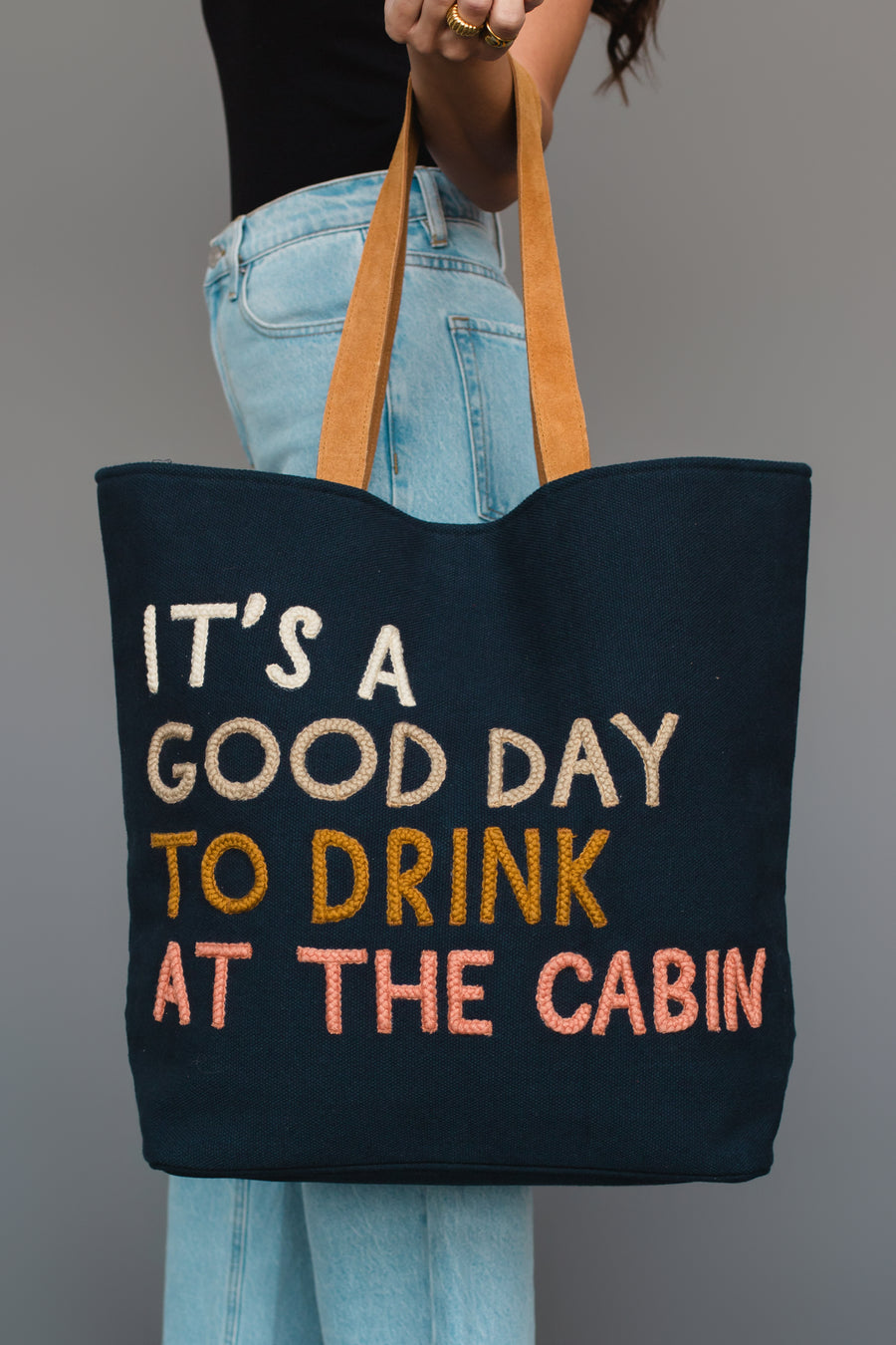 A person holds the Mustard & Pink Drink At The Cabin Tote by Panache Apparel Collection, featuring tan handles and text that reads, “ITS A GOOD DAY TO DRINK AT THE CABIN.” They are wearing light blue jeans and a black top.