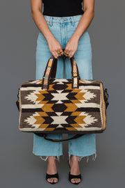 A person in a black top and light blue jeans holds the Panache Apparel Collection “Dark Gray, Camel & Black Western Duffel.” The person’s face is not visible.