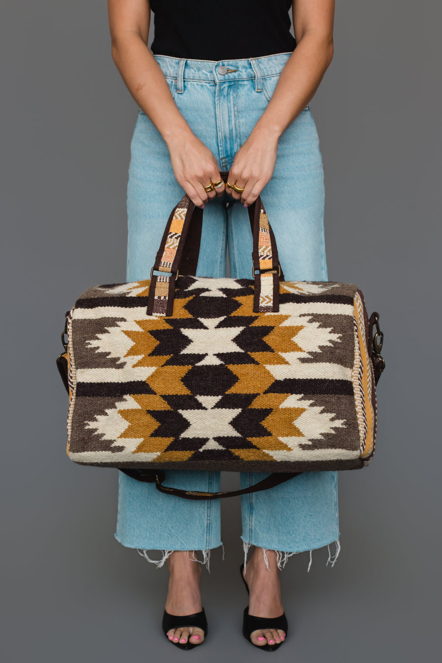 A person in a black top and light blue jeans holds the Panache Apparel Collection “Dark Gray, Camel & Black Western Duffel.” The person’s face is not visible.