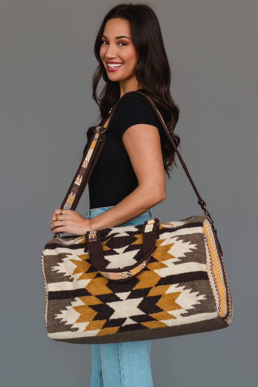 A woman smiles, wearing a black top and light blue jeans, as she holds the Panache Apparel Collection’s Dark Gray, Camel & Black Western Duffel, with its striking geometric pattern and brown strap, over her shoulder against a gray background.
