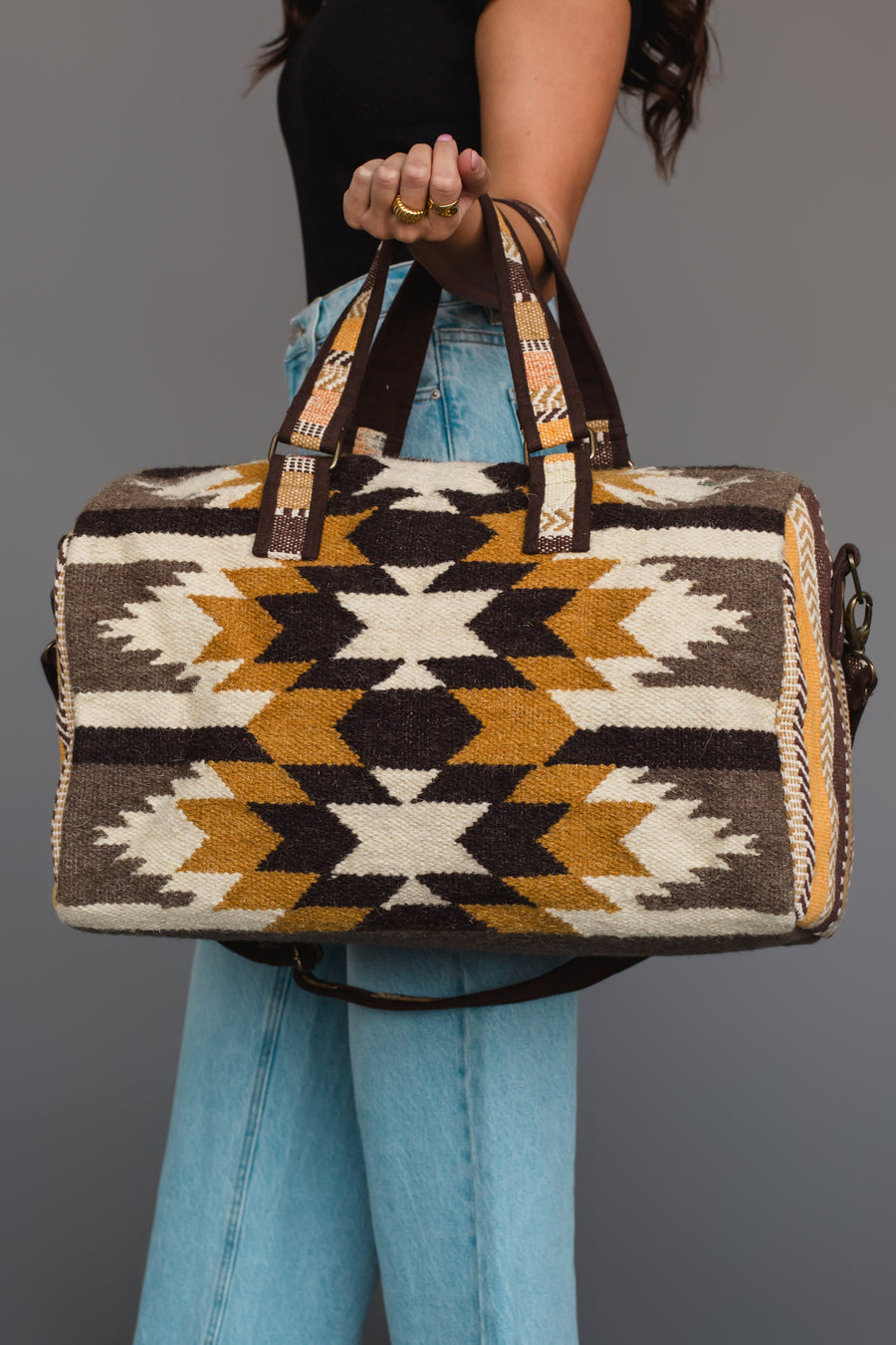 A person wearing a black top and blue jeans holds the Panache Apparel Collection Dark Gray, Camel & Black Western Duffel.