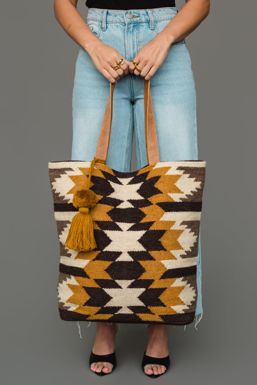 A person in light blue jeans and black sandals holds the Panache Apparel Collection’s Dark Gray, Camel & Black Western Tote against a gray background.