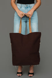 A person wearing a black top, blue jeans, and black sandals holds the Panache Apparel Collection Dark Gray, Camel & Black Western Tote against a gray background.