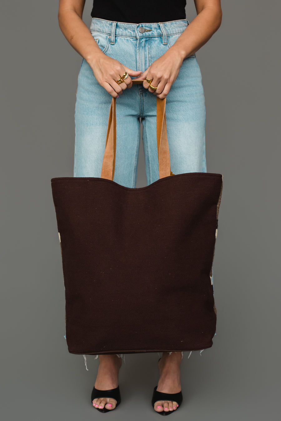 A person wearing a black top, blue jeans, and black sandals holds the Panache Apparel Collection Dark Gray, Camel & Black Western Tote against a gray background.