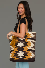 A woman with long brown hair smiles while carrying the Panache Apparel Collection’s Dark Gray, Camel & Black Western Tote, which features bold geometric patterns and a tassel. She pairs it with a black shirt and light jeans against a plain background.