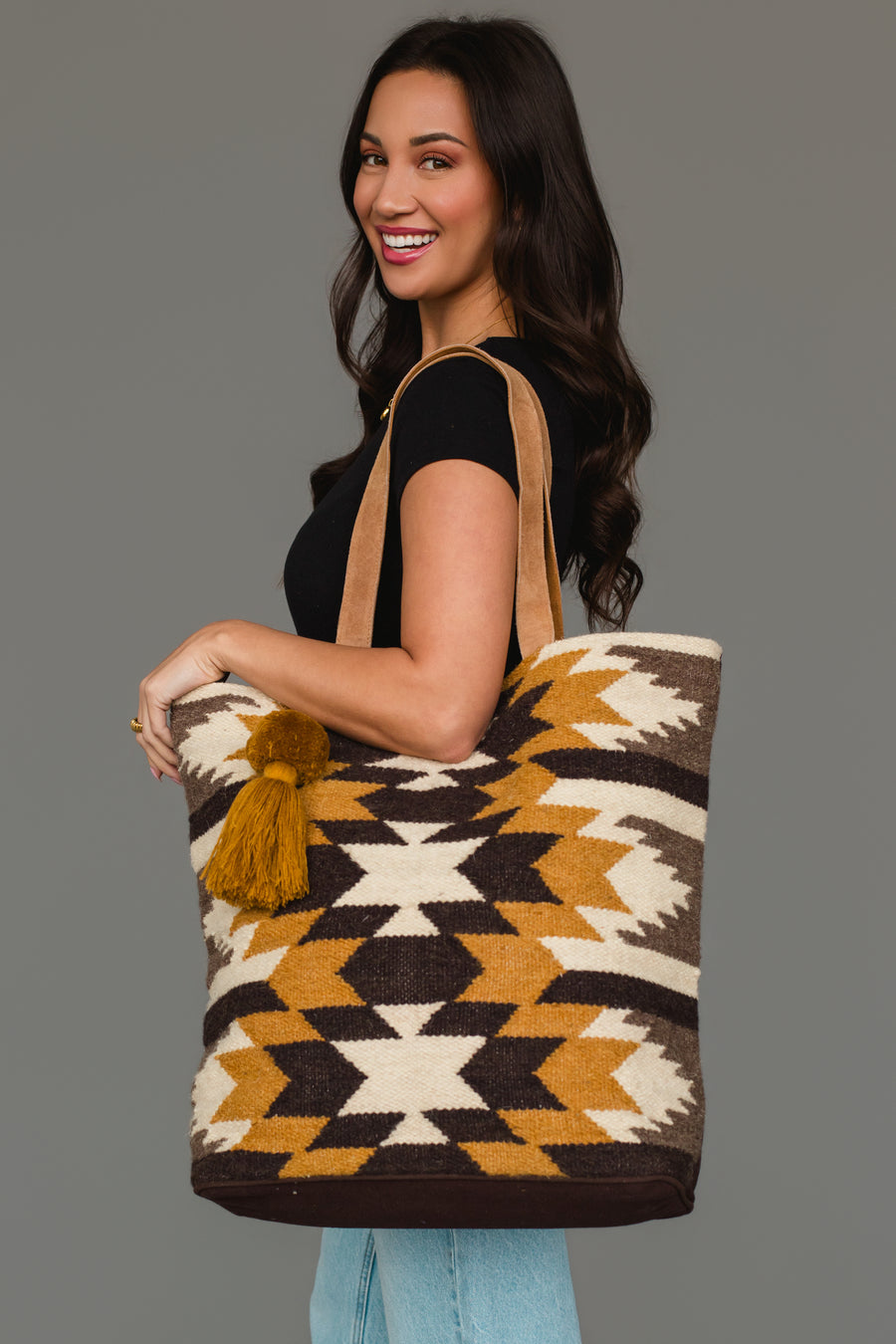 A woman with long brown hair smiles while carrying the Panache Apparel Collection’s Dark Gray, Camel & Black Western Tote, which features bold geometric patterns and a tassel. She pairs it with a black shirt and light jeans against a plain background.