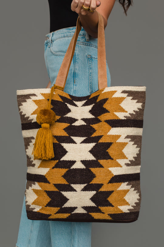 A person holds the Panache Apparel Collection Dark Gray, Camel & Black Western Tote, which features geometric patterns in dark gray, camel, and black with a leather strap and a tassel accent. The individual is wearing light blue jeans and a black top.