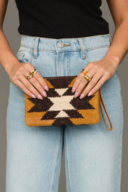 A person wearing light blue jeans and a black top holds the Camel & Black Western Wristlet from Panache Apparel Collection, a small clutch featuring a striking geometric pattern in black, white, and brown. Their manicured nails and multiple gold rings complete the look.