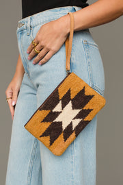 A person wearing light blue jeans holds the Camel & Black Western Wristlet by Panache Apparel Collection, a tan woven clutch featuring a black and white geometric pattern and brown strap. The person is also wearing gold rings.