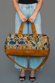 A person wearing light blue jeans and black heels holds the Panache Apparel Collection Mustard & Multicolored Patterned Duffel, featuring vibrant floral designs and brown corduroy accents with a long strap, against a gray background.