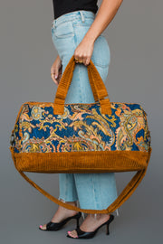 A person in light blue jeans holds the Mustard & Multicolored Patterned Duffel from Panache Apparel Collection, featuring brown corduroy accents and a shoulder strap, against a plain gray background.