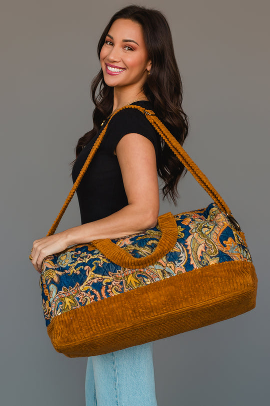 A smiling woman in a black top and jeans holds the Mustard & Multicolored Patterned Duffel from Panache Apparel Collection, featuring vibrant paisley designs and mustard yellow accents and straps, while standing against a plain gray background.
