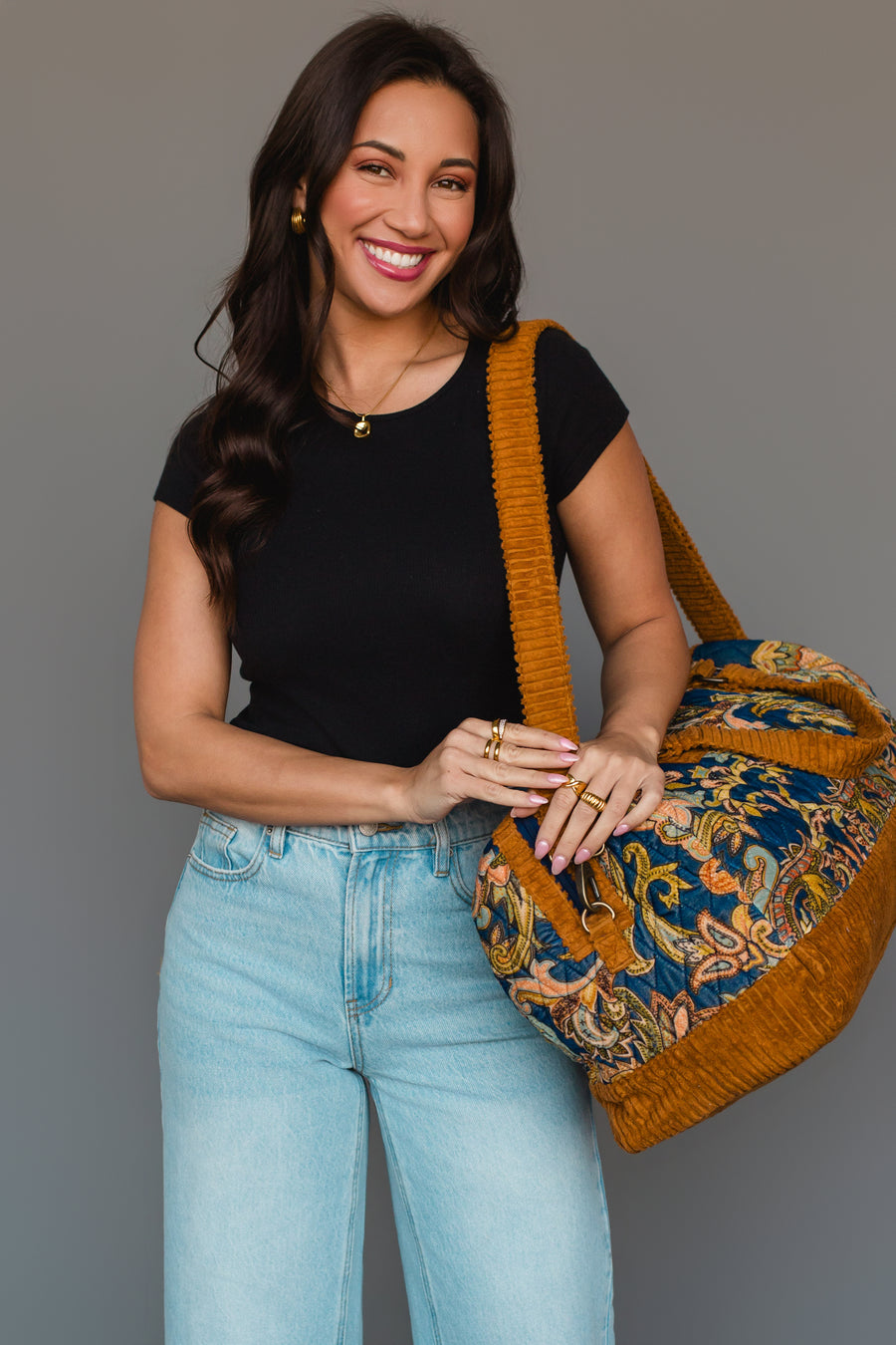 A woman with long dark hair, dressed in a black shirt and light blue jeans, smiles while holding the Panache Apparel Collection Mustard & Multicolored Patterned Duffel, which features vibrant designs and mustard yellow straps, standing against a plain gray background.
