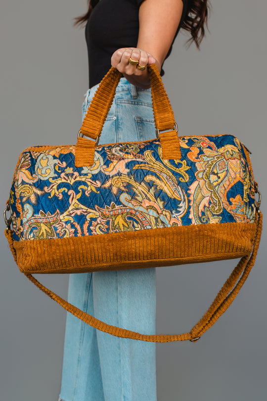 A woman in a black top and jeans holds the Panache Apparel Collection Mustard & Multicolored Patterned Duffel, featuring vibrant paisley designs with mustard yellow corduroy trim and handles, against a plain gray background.