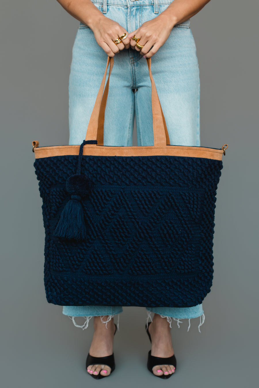 A woman carries the Panache Apparel Collection Navy Diamond Pattern Textured Tote, featuring tan leather handles and a tassel accent, styled with light wash jeans and black heeled sandals.