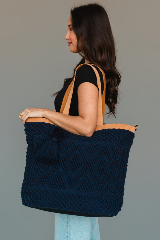 A woman with long dark hair in a black top and light jeans holds the Panache Apparel Collection Navy Diamond Pattern Textured Tote, featuring tan straps, over her shoulder while standing against a plain gray background.