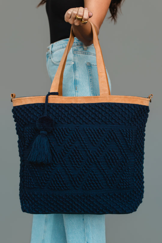 A woman in jeans and a black top holds the Panache Apparel Collection Navy Diamond Pattern Textured Tote, featuring tan handles and a decorative tassel, against a plain gray background.