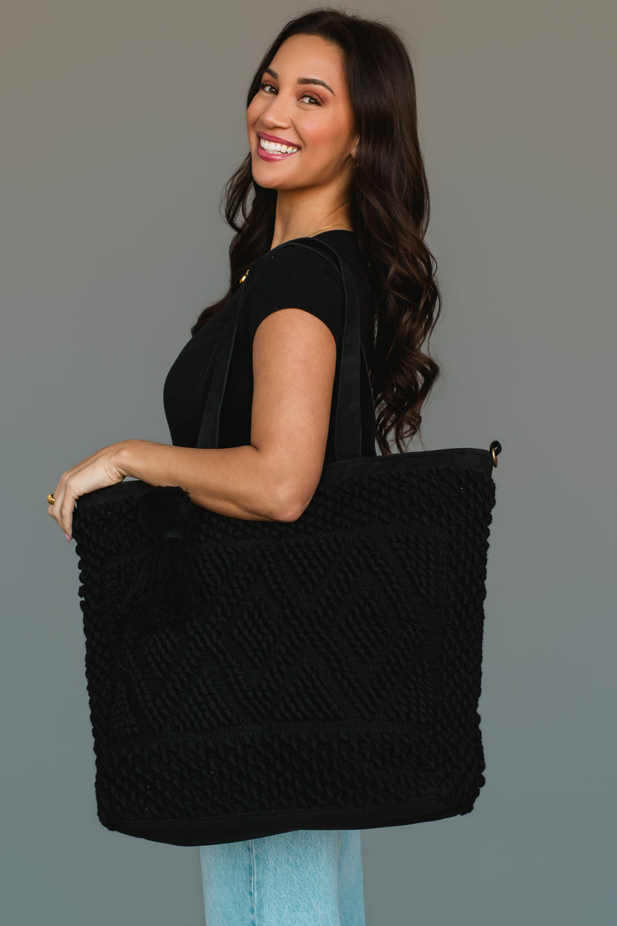 A woman with long dark hair, dressed in a black top and light blue jeans, smiles as she holds the Panache Apparel Collection Black Diamond Pattern Textured Tote over her shoulder against a plain gray background.