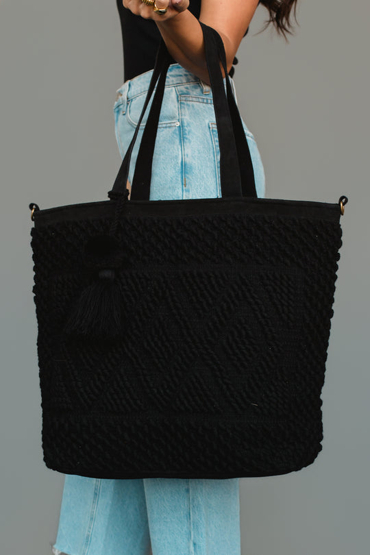 A woman in light blue jeans and a black top holds the Panache Apparel Collection Black Diamond Pattern Textured Tote, featuring tassel detailing and matching black straps.