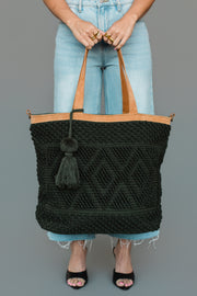 A person in jeans and black heels holds the Panache Apparel Collection’s Dark Green Diamond Pattern Textured Tote, featuring tan handles, a tassel, and geometric designs. Only the lower body and hands are visible against a plain gray background.