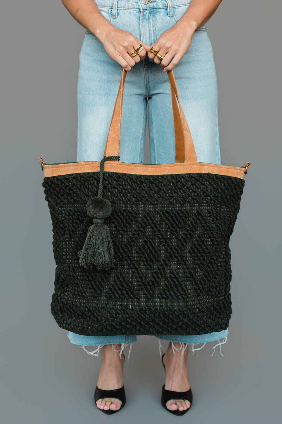 A person in jeans and black heels holds the Panache Apparel Collection’s Dark Green Diamond Pattern Textured Tote, featuring tan handles, a tassel, and geometric designs. Only the lower body and hands are visible against a plain gray background.