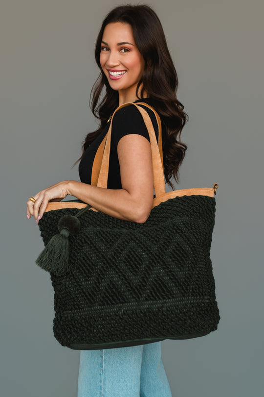 Smiling woman with long dark hair wearing a black shirt and light blue jeans, holding the Panache Apparel Collection Dark Green Diamond Pattern Textured Tote, which features camel-colored straps and a tassel, standing against a plain gray background.
