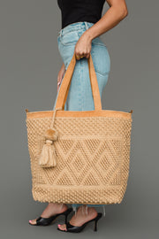 A woman wearing a black top, blue jeans, and black heels holds the panacheaccessories Tan Diamond Pattern Textured Tote an elegant tan bag with leather handles and a decorative tassel against a gray background. The tote’s sophisticated diamond pattern adds subtle flair to her look.