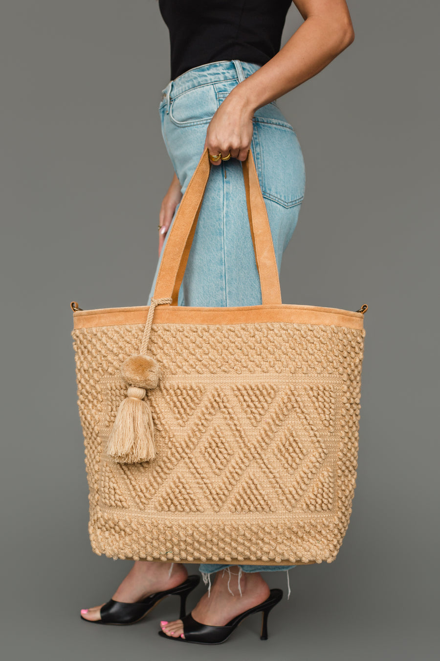 A woman wearing a black top, blue jeans, and black heels holds the panacheaccessories Tan Diamond Pattern Textured Tote an elegant tan bag with leather handles and a decorative tassel against a gray background. The tote’s sophisticated diamond pattern adds subtle flair to her look.