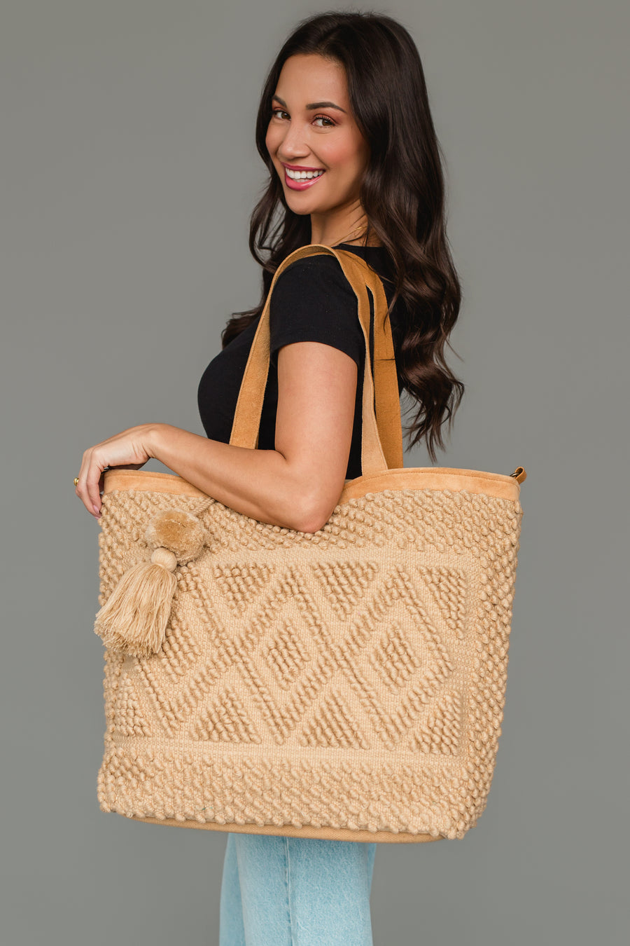 A woman smiles while wearing a black shirt and light jeans, holding the panacheaccessories Tan Diamond Pattern Textured Tote with leather shoulder straps and tassel accents, standing against a plain gray background.